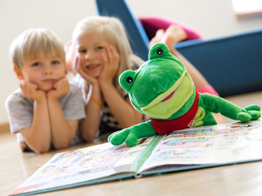 Dieses Bild zeigt zwei Kinder auf die Ellenbogen gestützt vor einem Buch liegen. Außerdem im Bild ein grüner Plüschfrosch.