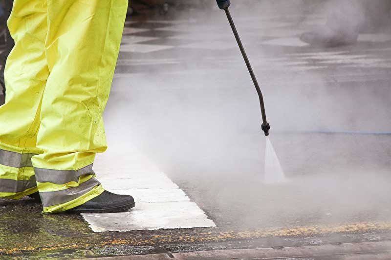Das Bild zeigt zwei gelbe Hosenbeine und die Düse eines Dampfstrahlers welcher die Straße reinigt.