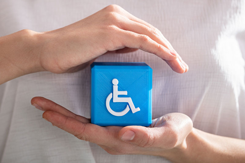 Dieses Bild zeigt eine Hand welche einen blauen Würfel mit dem Rollstuhlfahrersymbol hält. Die andere Hand wird schützend wie ein Dach darüber gehalten.