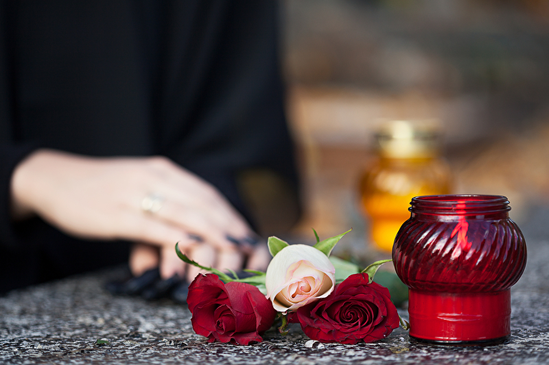 Dieses Bild zeigt eine Hand auf einem Grabstein liegend und eine Rose haltend. Danebei steht eine Kerze.