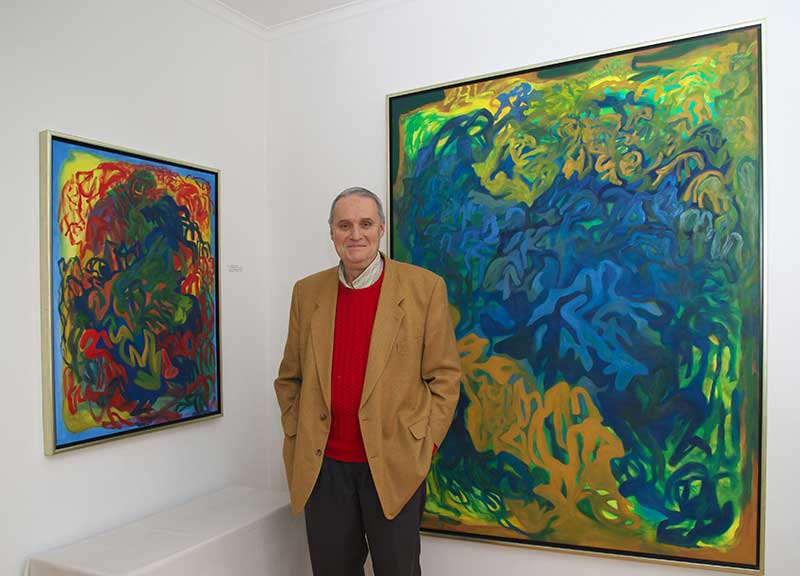 Das Bild zeigt den Künstler Karl Aichinger vor zwei seiner Gemälde stehend.